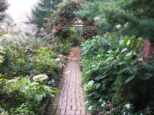 A lush garden path lined with vibrant flowers and greenery, leading to a shaded archway.