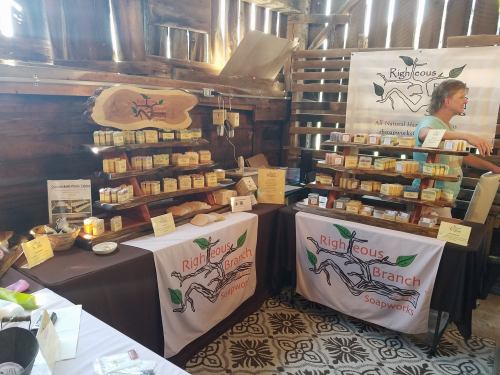 A rustic market display featuring handmade soaps on wooden shelves and tables, with a vendor in the background.