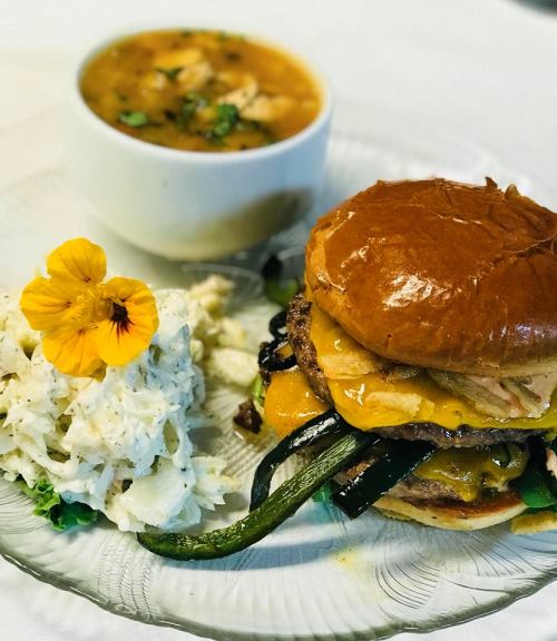 A delicious burger with cheese and grilled vegetables, served with coleslaw and a bowl of soup, garnished with a flower.