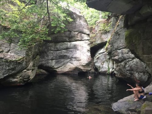 A serene natural pool surrounded by large rocks and trees, with a person swimming and another sitting on the edge.