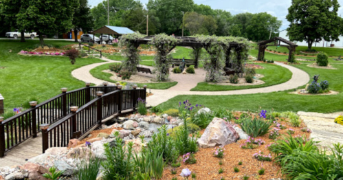 A scenic garden with winding paths, wooden bridges, and lush greenery, featuring flower beds and decorative arches.