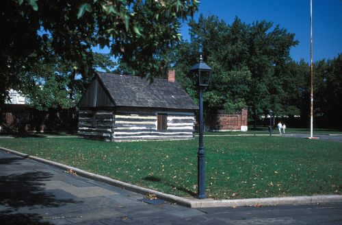 A small log cabin surrounded by green grass and trees, with a lamp post nearby and a clear blue sky.