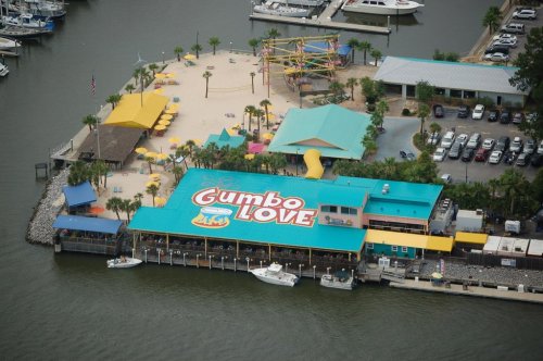 Aerial view of Gumbo Love restaurant with colorful roofs, beach area, and boats docked nearby.