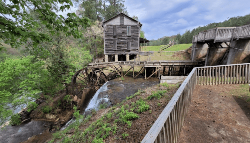 A historic watermill with a wooden structure and a large waterwheel, surrounded by greenery and a flowing stream.