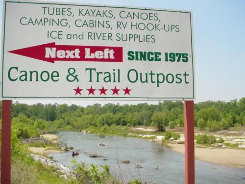 Sign for "Canoe & Trail Outpost" with services like tubing, kayaking, and camping, next to a river.