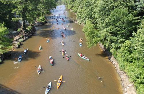 Aerial view of a river with numerous kayakers and canoers enjoying a sunny day surrounded by lush greenery.