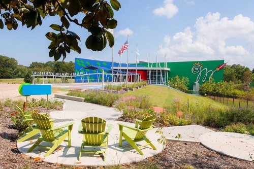 Colorful modern building with flags, surrounded by greenery and outdoor seating in a sunny landscape.