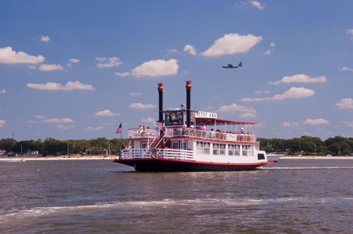 A paddleboat with two smokestacks sails on a river under a blue sky with fluffy clouds, while an airplane flies above.