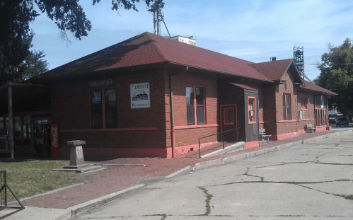 Historic brick train depot with a red roof, featuring windows and signage, set against a clear blue sky.