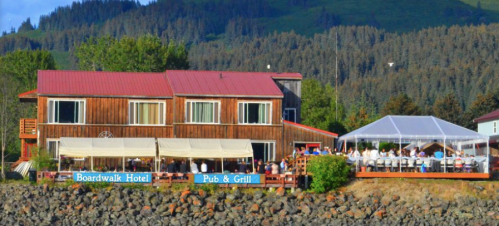 A wooden hotel with a red roof and outdoor dining area by the water, surrounded by trees and mountains in the background.
