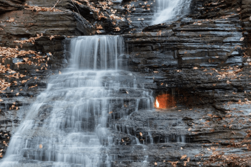 A serene waterfall cascading over dark rocks, surrounded by autumn leaves.