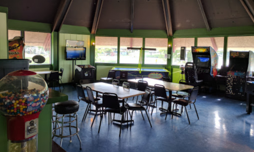 A colorful arcade room with tables, chairs, arcade machines, and a gumball machine in a bright, inviting space.