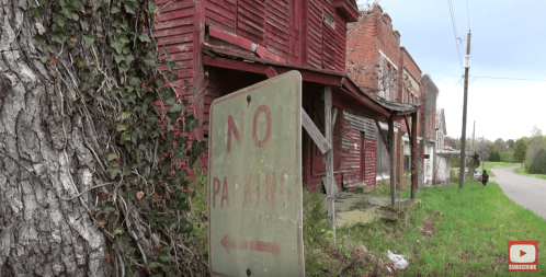 An old "No Parking" sign next to a weathered building, surrounded by overgrown grass and trees.