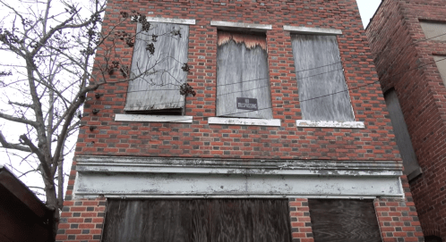 Abandoned brick building with boarded windows and peeling paint, surrounded by bare trees.