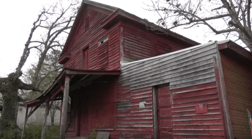 A weathered red house with peeling paint, surrounded by bare trees and a cloudy sky.
