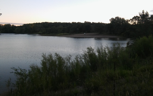 A serene lakeside view at dusk, with trees lining the shore and a sandy beach in the distance.