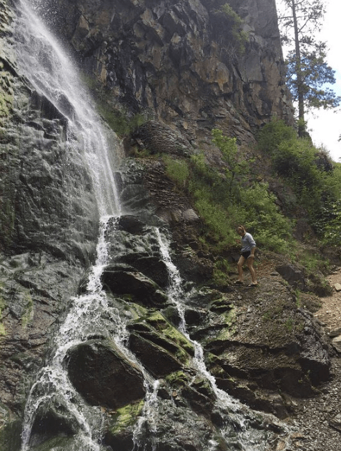 A person stands near a waterfall cascading down rocky terrain surrounded by greenery.