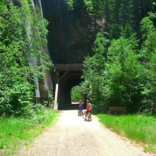 Two cyclists ride on a dirt path through a forested area, approaching a large tunnel surrounded by greenery.