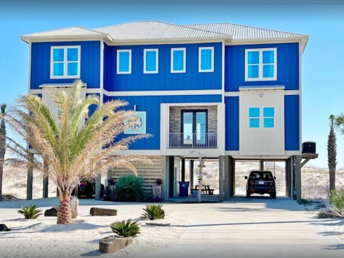 A large blue beach house with palm trees and a driveway, set against a sandy landscape.