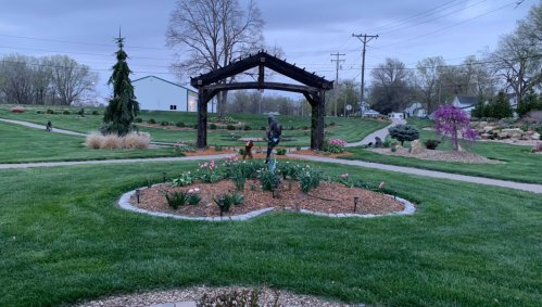 A landscaped garden with a wooden arch, flower beds, and green grass under a cloudy sky.