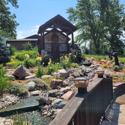 A scenic garden with a stone cottage, vibrant flowers, and a small pond, framed by trees and a wooden walkway.