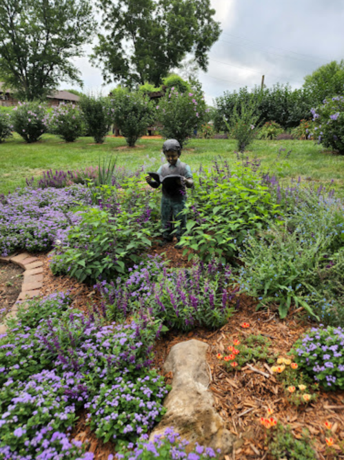 A statue of a child reading surrounded by vibrant purple and colorful flowers in a lush garden setting.