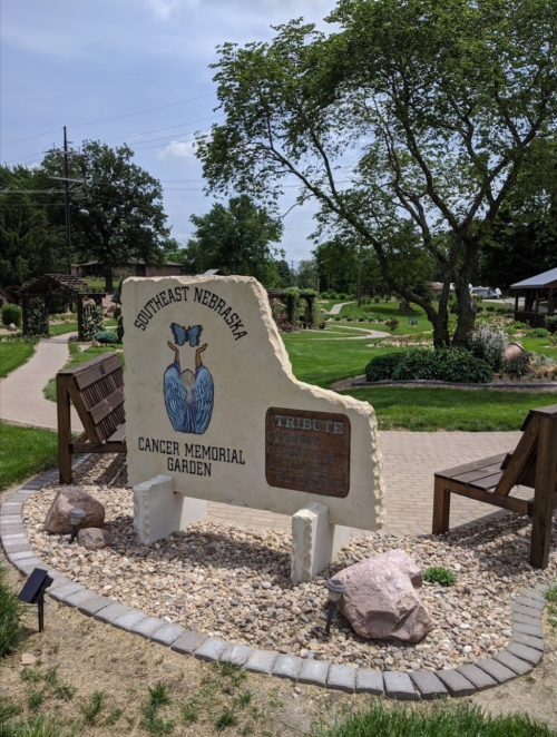 A stone monument in a garden, reading "Southeast Nebraska Cancer Memorial Garden," surrounded by benches and greenery.