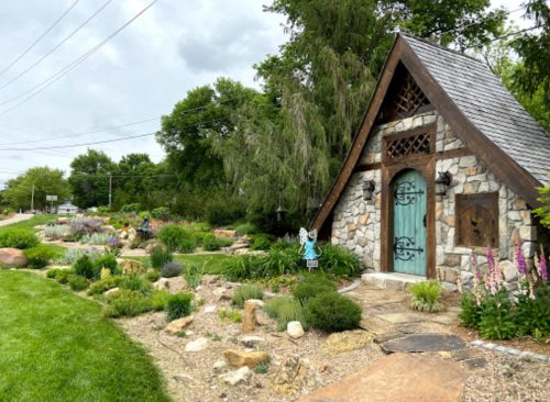 A charming stone cottage with a teal door surrounded by a lush garden and greenery on a cloudy day.