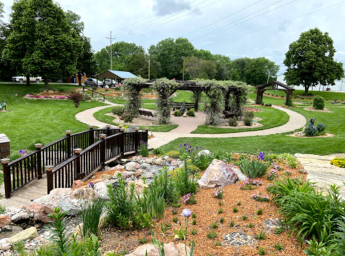 A landscaped park featuring a winding path, wooden bridge, and flower beds with greenery and stone accents.