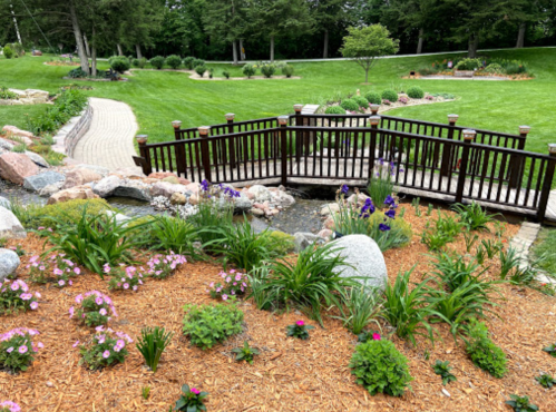 A serene garden scene featuring a wooden bridge over a stream, surrounded by colorful flowers and lush greenery.