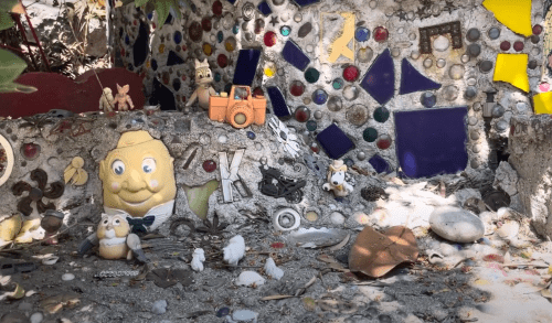 A colorful mosaic wall adorned with various quirky objects and figurines, set in a whimsical outdoor space.