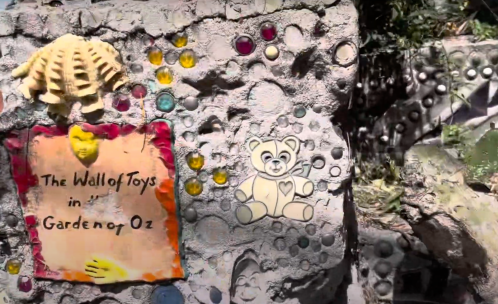Colorful wall with a sign reading "The Wall of Toys in Garden of Oz," featuring a teddy bear and decorative stones.