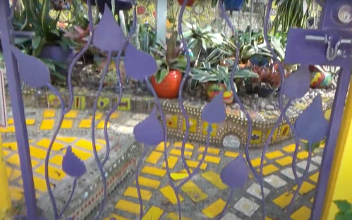 Colorful mosaic garden with vibrant plants, decorative pathways, and artistic metalwork in the foreground.
