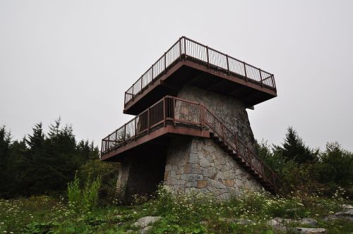 A two-level observation tower made of stone and wood, surrounded by greenery and a cloudy sky.