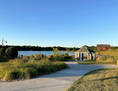 A serene park scene featuring a lake, gazebo, and walking path surrounded by lush greenery under a clear blue sky.