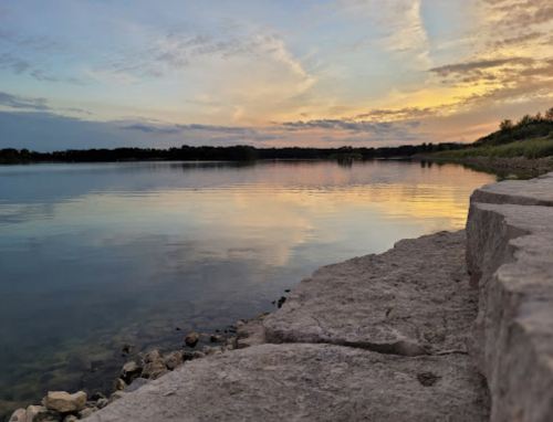 A serene lakeside view at sunset, with calm waters reflecting colorful clouds and a rocky shoreline.