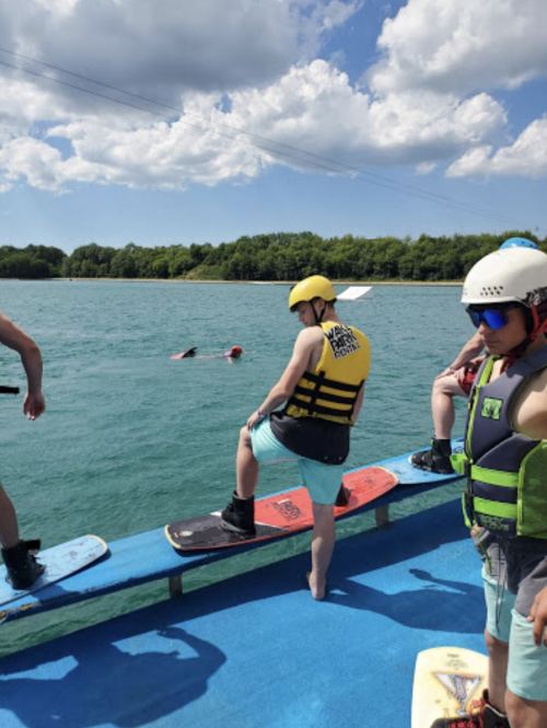 A group of people on a boat preparing to wakeboard, with one person in the water and a sunny sky above.