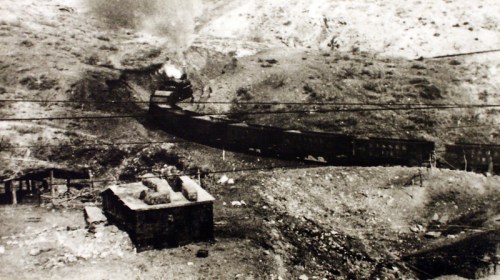 A vintage black-and-white photo of a steam train winding through a mountainous landscape with smoke billowing.