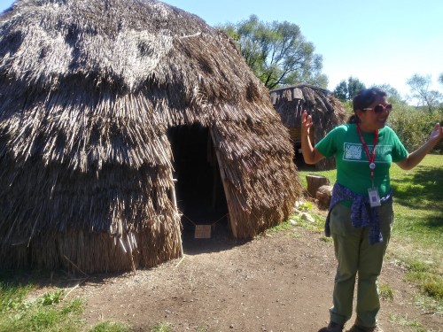 A person in a green shirt gestures near a straw-covered hut in a grassy area, with trees in the background.