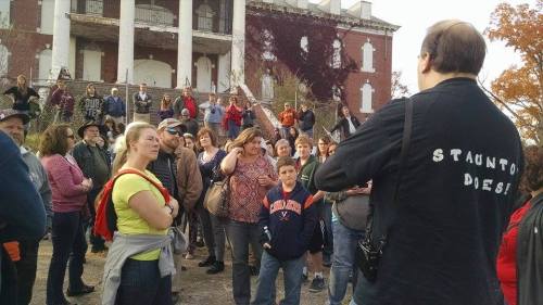 A crowd gathers outside a historic building, listening to a speaker wearing a shirt that says "STAUNTON DIES!"