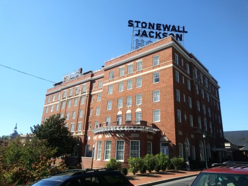 Historic Stonewall Jackson Hotel, a brick building with a prominent sign, surrounded by greenery and a clear blue sky.