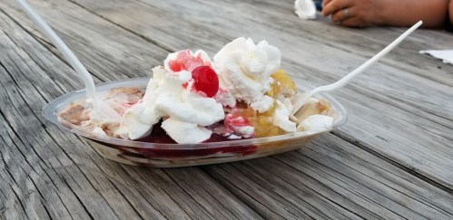 A dessert bowl filled with ice cream, whipped cream, cherries, and syrup, sitting on a wooden table.