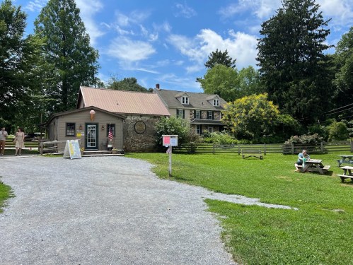 A quaint building with a stone facade and a red roof, surrounded by greenery and picnic tables on a sunny day.
