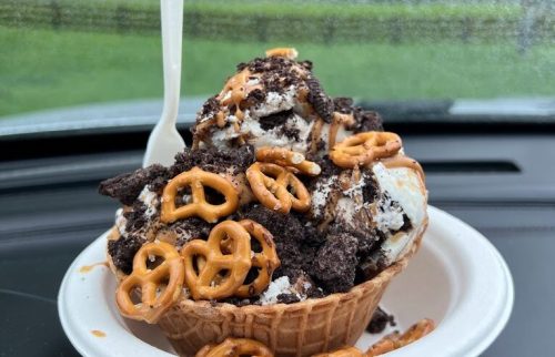A waffle cone filled with ice cream, crushed Oreos, and pretzels, sitting on a car dashboard with a rainy window in the background.