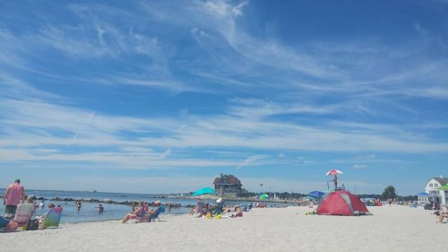 A sunny beach scene with people relaxing, colorful umbrellas, and a distant building under a blue sky with wispy clouds.