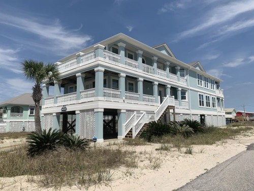 A large beach house with a light blue exterior, white railings, and palm trees, set against a sandy landscape.