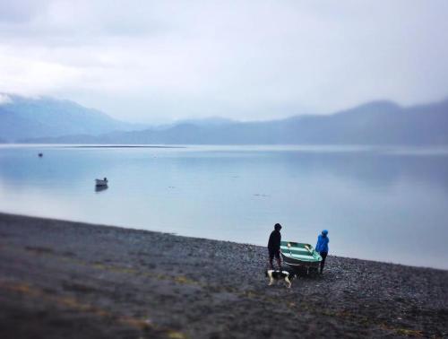 A person and a dog near a small boat on a calm, misty lake shore with mountains in the background.