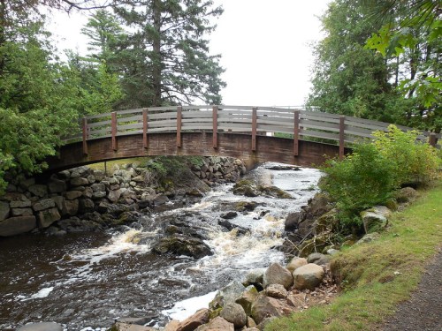 A wooden bridge arches over a flowing stream, surrounded by lush greenery and rocky banks.