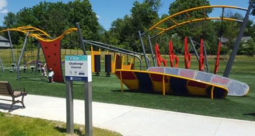 Colorful outdoor challenge course with climbing structures and obstacles, set in a grassy park area.