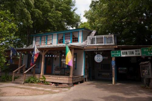 A colorful two-story building with flags, featuring a porch and a sign for a night club. Lush greenery surrounds it.